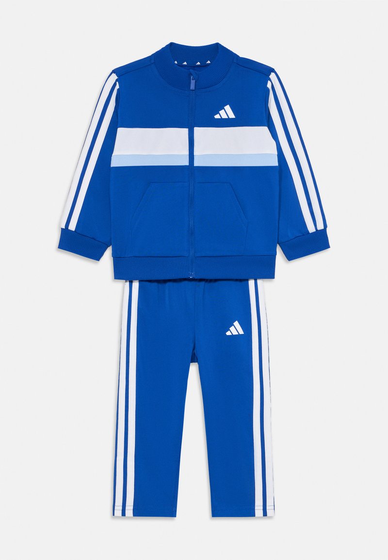 Kék sportos tréningruha, fehér és világoskék vízszintes csíkokkal ellátott zipzáros dzsekivel és hozzá illő nadrággal, mindkettő oldalsó csíkokkal.