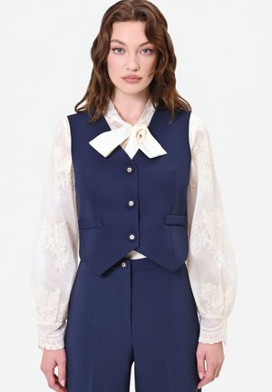 Donna con capelli castani mossi che indossa un gilet e pantaloni blu navy con una camicetta bianca in pizzo e papillon, in piedi davanti a uno sfondo chiaro uniforme.