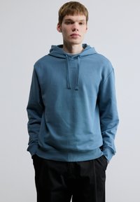 Sweatshirt com capuz em tecido azul claro macio, com bolso canguru frontal, punhos canelados e um pequeno logótipo no peito.
