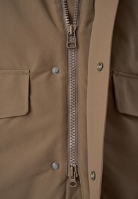 Brauner Jackenstoff mit glatter Textur, ausgestattet mit einem zentralen Reißverschluss und zwei vorderen Taschen mit Druckknopfverschlüssen. Details umfassen einen metallischen Reißverschluss-Anhänger.