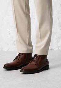 Brązowe skórzane botki do kostki z okrągłymi noskami, sznurówkami i przeszyciami, noszone z jasnobeżowymi spodniami na teksturowanym tle.