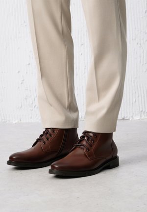 Brązowe skórzane botki do kostki z okrągłymi noskami, sznurówkami i przeszyciami, noszone z jasnobeżowymi spodniami na teksturowanym tle.