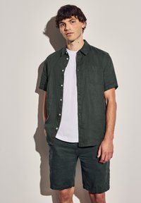 Mörkgrön kortärmad skjorta över en vit t-shirt, kombinerat med mörkgröna shorts. Skjortan har knappar och en bröstficka, med texturerad tyg.