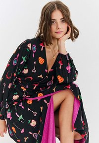 Czarna sukienka kopertowa z kolorowymi wzorami owoców, kluczy i luster. Posiada długie rękawy i różowy dół w kropki.