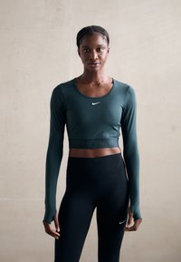 Cămașă verde cu mânecă lungă și guler rotund, cu logo-ul Nike, asortată cu colanți negri cu talie înaltă, cu o textură fină.