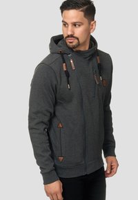 Sudadera gris con cremallera y cuello alto, con detalles en cuero marrón, cordones y bolsillos laterales triangulares. Tejido suave y texturizado.