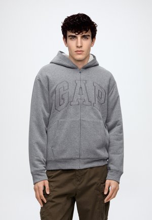 Jeune homme aux cheveux bouclés et foncés portant un sweat à capuche gris GAP zippé et un pantalon marron, debout devant un fond clair uni.