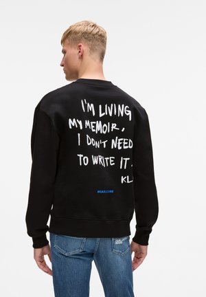 Sudadera negra con cuello redondo, que presenta texto manuscrito en blanco en la parte posterior. El texto incluye "#KARLCORE" en azul en la parte inferior.