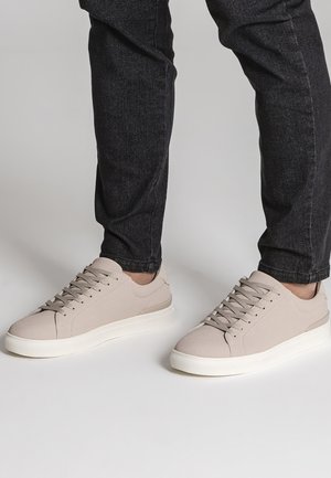 Person, die hellbeige Low-Top-Sneaker mit weißen Sohlen und schwarzen Jeans trägt, steht auf einer einfarbigen hellgrauen Oberfläche.