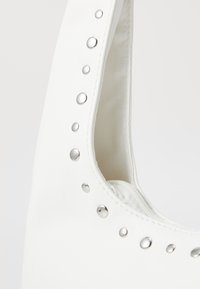 Sac à main blanc avec une surface texturée, comportant des clous en argent le long de l'ouverture. Fermeture éclair visible à l'intérieur. Design élégant et structuré.