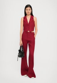 Rotes, kurzes ärmelloses Oberteil mit einer Schlitzkragen und vorderen Knöpfen, kombiniert mit roten, ausgestellten Hosen. Das Model hält eine schwarze Tasche mit Nieten.