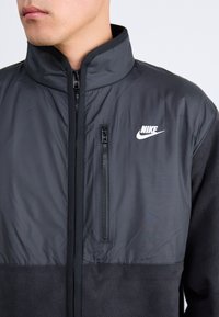 Moški nosi črno jakno Nike z visokim ovratnikom, sprednjim zadrgo in žepom na prsih.