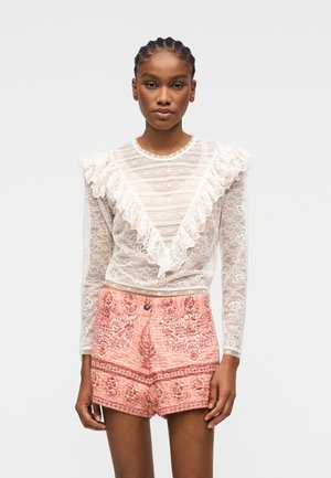 Frau trägt eine transparente weiße Spitzenbluse mit langen Ärmeln und Rüschen sowie hoch taillierte korallfarbene Shorts mit Muster, steht vor einem einfarbigen Hintergrund.