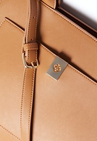 Bolso de mano de cuero marrón claro con textura suave, diseño geométrico, correa ajustable y un acento decorativo de hardware plateado.