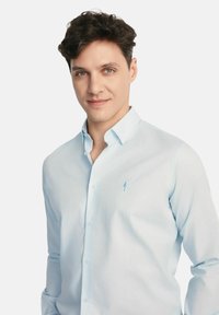 Jasnoniebieska koszula z guzikami, uszyta z tkaniny o gładkiej fakturze, z małym haftowanym logo na lewej kieszeni piersiowej.