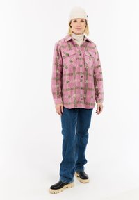 Vrouw in een button-up jack met een roze en groene print, een beige gebreide beanie, een beige coltrui, blauwe jeans en zwarte laarzen met beige zolen.