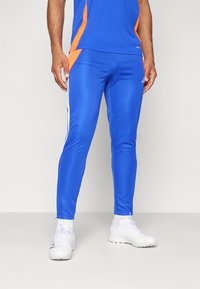Blauwe sportbroek met oranje accentpanelen, getailleerde pasvorm en witte zijstrepen. Gemaakt van glanzend, soepel materiaal. Gecombineerd met witte schoenen.