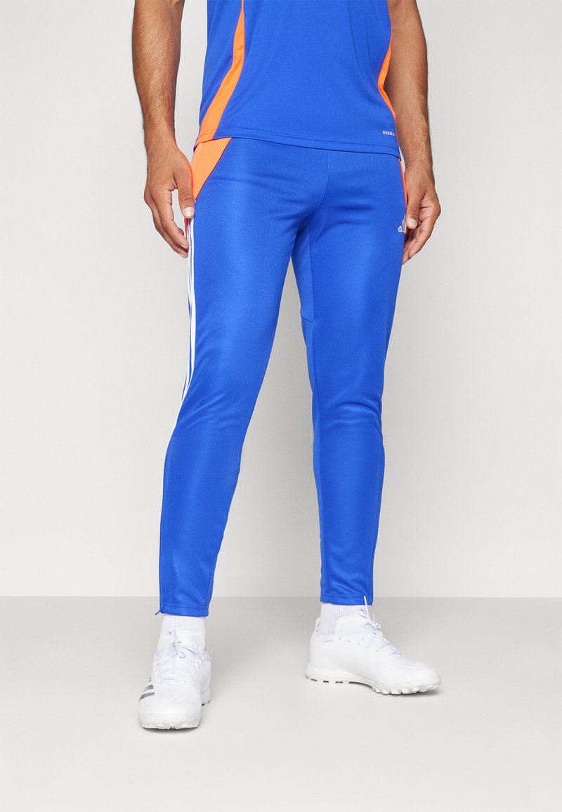 Blauwe sportbroek met oranje accentpanelen, getailleerde pasvorm en witte zijstrepen. Gemaakt van glanzend, soepel materiaal. Gecombineerd met witte schoenen.