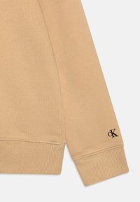 Manica di felpa in cotone beige con polsino a costine e logo "cK" nero vicino al polso. Texture liscia con cuciture precise.