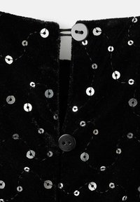 Černá tkanina s jemnou texturou, zdobená metalickými flitry uspořádanými do vířivého vzoru, zapnutá černým knoflíkem na výstřihu.