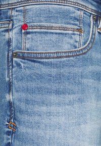 Light blue denim jeans with a front pocket, orange stitching, and a red button accent. Texture appears smooth with a slight fade.