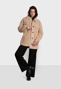 Casaco de pele falsa em bege com fecho frontal por botões, duas bolsas no peito e gola, combinado com calças largas pretas e sapatos pretos.