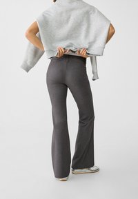 Pantalons gris évasés en tissu texturé, taille haute, avec poches arrière, associés à un pull gris clair drapé sur les épaules.