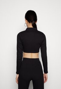 Top cropped preto com textura canelada, mangas longas e design com colarinho. Apresenta um decote alto e um corte que expõe a barriga.