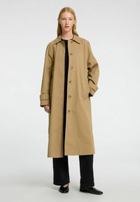 Beige lang frakke lavet af glat stof, med en klassisk krave, knaplukning og sidelommer. Båret over en sort top og bukser.