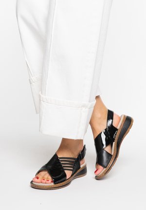Feet wearing black strappy sandals with low wedge heels and painted toenails, paired with white cuffed pants against a plain background.