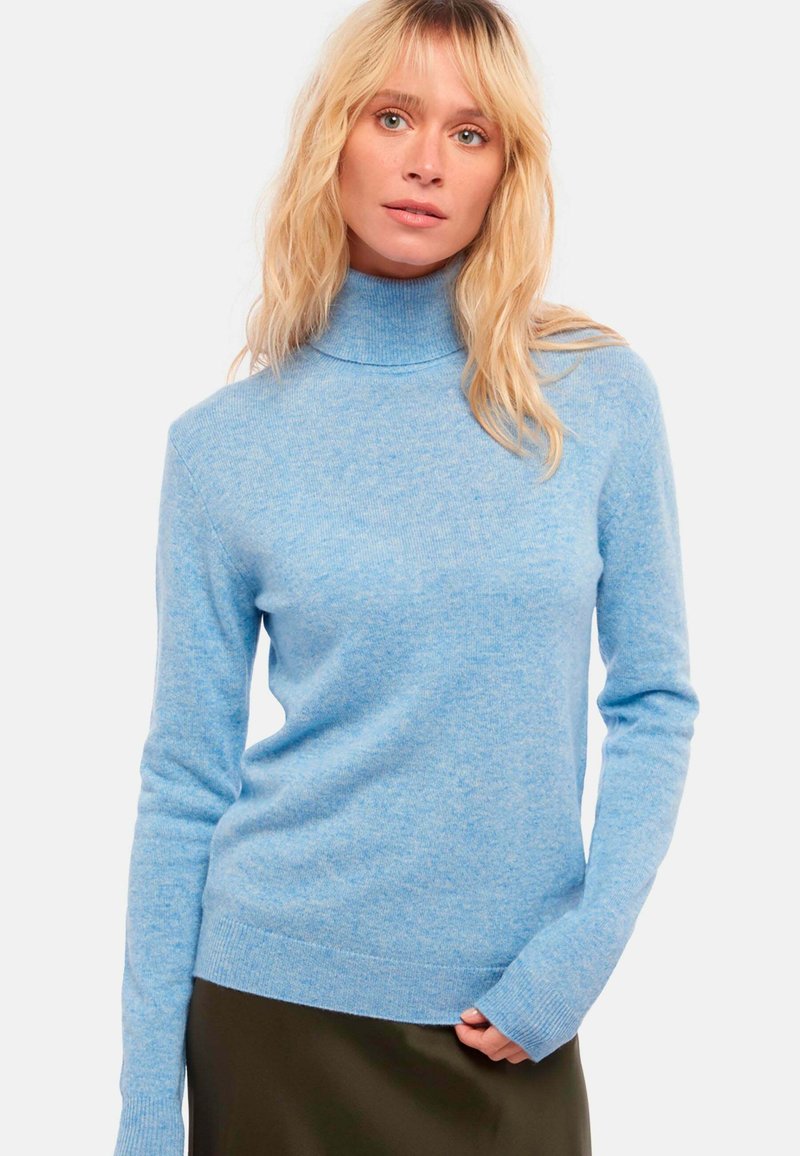 Un pull à col roulé bleu clair en matière tricotée douce, avec des manches longues et un ourlet côtelé, porté sur une jupe vert foncé.