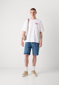 Witte katoenen T-shirt met rode "Calvin Klein" logo, blauwe denim shorts, beige sneakers en een lichtbeige schoudertas. Korte, rechte stijl.