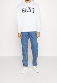 Sudadera blanca con "GANT" en letras negras en negrita, combinada con vaqueros rectos de color azul claro y zapatillas blancas, de pie contra un fondo liso.