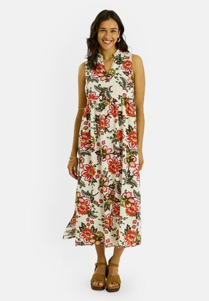 Mujer sonriente que lleva un vestido midi blanco sin mangas con un gran estampado floral rojo y rosa y sandalias marrones de plataforma.