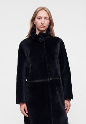 Femme blonde portant un manteau noir à col haut et en peluche, debout devant un fond gris clair uni.