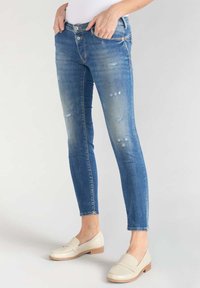Vaqueros ajustados de mezclilla en un tono azul, con detalles desgastados y cinco bolsillos. Combinados con unos mocasines de color claro.