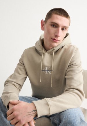 Beige hoodie met trekkoorden, met een logo op de borst. Draag met lichtblauwe jeans, die een gladde stoftextuur laten zien.