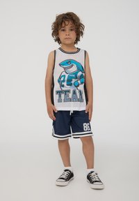 Canotta bianca in rete con grafiche di uno squalo blu, abbinata a pantaloni sportivi navy con accenti bianchi e grande stampa "86". Sneaker nere.