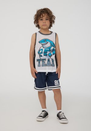 Canotta bianca in rete con grafiche di uno squalo blu, abbinata a pantaloni sportivi navy con accenti bianchi e grande stampa "86". Sneaker nere.