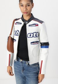 Casaquinho de motoqueiro em pele branca com painéis em malha, detalhes em azul e vermelho, fecho com zip e patches bordados. Combinado com calças de ganga.