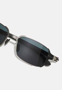 Γυαλιά ηλίου με σκούρους φακούς, μεταλλικό σκελετό, γωνιώδη σχεδίαση και ρυθμιζόμενα μαξιλαράκια μύτης. Ο σκελετός έχει δίχρωμο σχέδιο σε χρυσό και μαύρο.