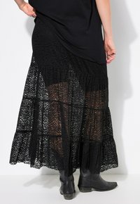 Schwarzer Spitzen-Maxi-Rock mit gestuftem Design, strukturiertem Muster und transparenter Stoff, kombiniert mit schwarzen Lederstiefeln.