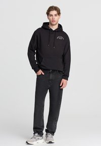 Czarna bluza z kapturem z przednią kieszenią typu kangur, z małym białym wzorem tekstowym. Nosi ją osoba w czarnych dżinsach i szarych trampkach. Proste tło.