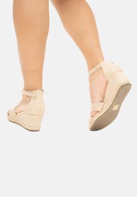 Sandali con zeppa in suede beige, con punta aperta, cinturino alla caviglia e dettaglio in juta intrecciata. Texture liscia, tacco elevato e design minimalista.