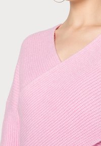 Kobieta ubrana w miękki, różowy, prążkowany sweter z dekoltem w serek i kopertowym przodem, widoczna od szyi do połowy tułowia.