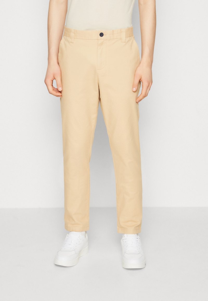 Personne portant un pantalon beige à jambes droites et des baskets blanches, debout devant un fond clair uni.