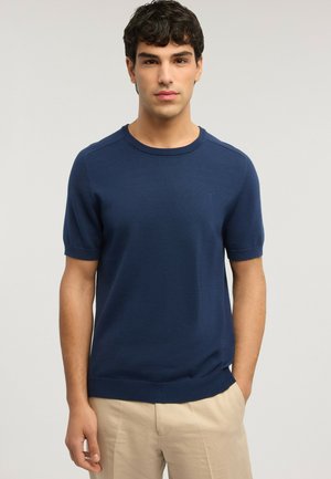 Uomo con indosso una maglietta a maniche corte blu navy in maglia e pantaloni beige, in piedi davanti a uno sfondo chiaro uniforme.