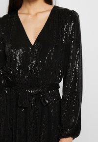 Robe noire en sequins avec un décolleté en V profond, de longues manches bouffantes et une taille nouée. Le tissu est texturé, avec des sequins brillants et réfléchissants.