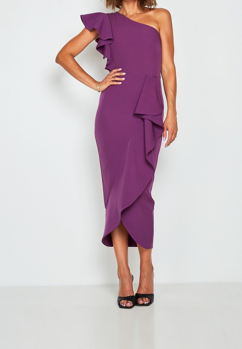 Femme portant une robe midi violette à une épaule avec des détails à volants et des talons hauts noirs ouverts, debout devant un fond blanc uni.