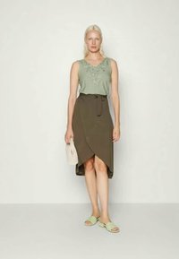 Top sem mangas verde-claro com decote bordado, combinado com uma saia envolvente verde-oliva que se ata na cintura. Modelo usa chinelos verde-menta.